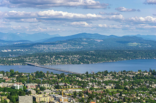 The image shows a picturesque view of a city with a large body of water, possibly a lake or river, surrounded by mountains and lush greenery under a partly cloudy sky.