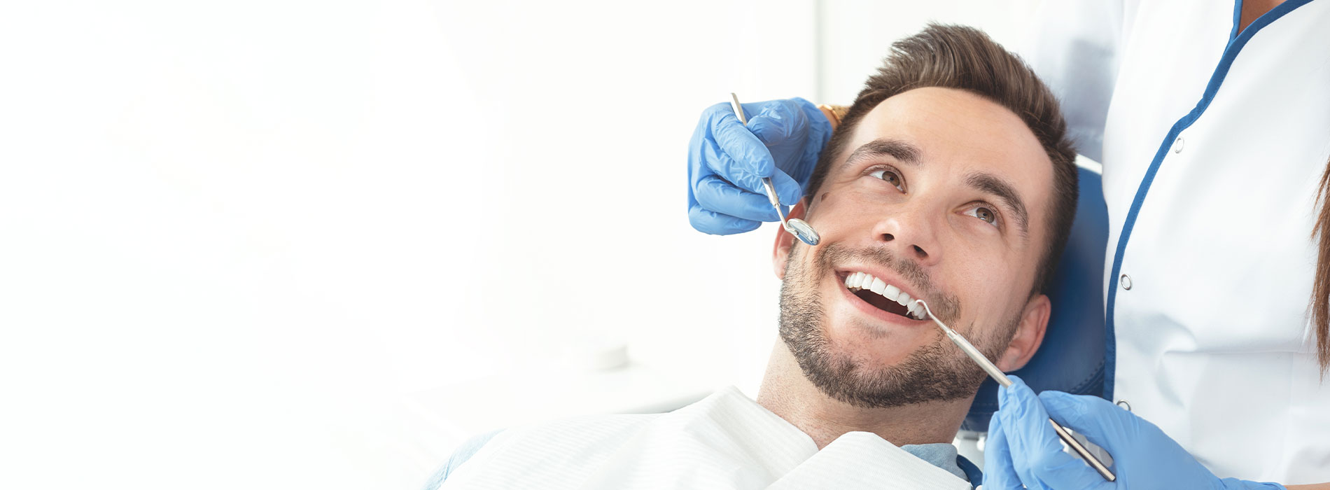 The image shows a man lying on a dental chair with a smile on his face, receiving dental care from a dentist who is wearing protective gear.
