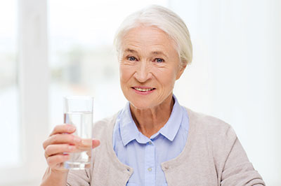 An elderly woman holding a glass of water with both hands, smiling at the camera.