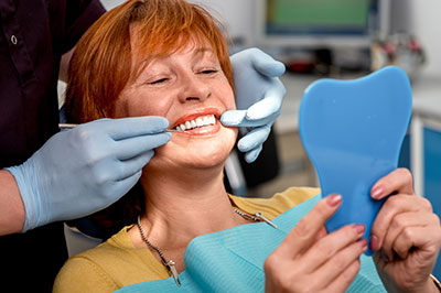 This image shows a woman sitting in a dental chair, holding up a blue dental model with a smile on her face, while a dentist examines her teeth. The setting suggests a dental office environment.