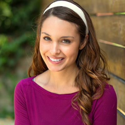 A young woman with long brown hair, wearing a purple top, poses for a portrait with a smile, against a wooden fence background.