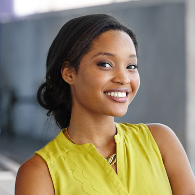 A woman with a bright smile poses confidently against a backdrop, wearing a yellow top and black pants.