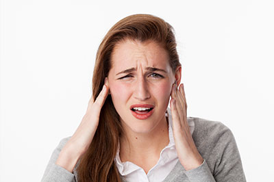 The image shows a young woman with her hand on her head, appearing to be experiencing discomfort or pain, possibly due to an ear issue, wearing a gray sweater, with a neutral background.