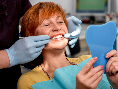 A woman with red hair is seated in a dental chair, smiling at the camera while holding up a blue tooth-shaped object, with dental tools around her mouth.