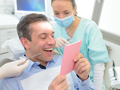The image shows a man sitting in a dental chair with a big smile, holding up a pink card with both hands while looking at it, surrounded by dental equipment and a smiling dental professional wearing a surgical mask and gown.