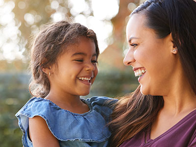 The image shows a woman with a child, both smiling and looking at each other. They are outdoors during daylight.