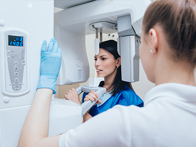 The image shows two individuals, likely medical professionals, standing in front of a large piece of medical imaging equipment, possibly an MRI machine, with one person looking at the equipment screen while another looks on.