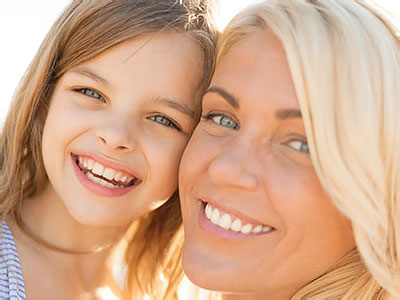 The image shows a woman with a child, both smiling at the camera, against a blurred background suggesting a sunny outdoor setting.