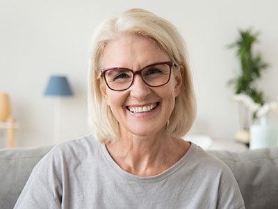 The image features a woman with short blonde hair smiling at the camera, wearing glasses and a gray top, seated indoors against a white wall with a lamp visible behind her.