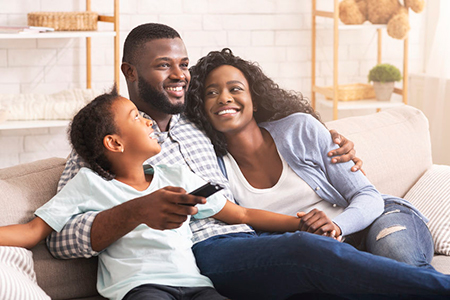 A family of four sitting on a couch, with an adult holding two children close, smiling and looking at something off-camera.