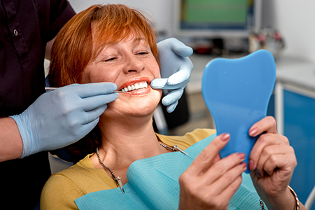 A woman sitting in a dental chair with a blue mouthguard on her teeth, smiling at the camera while holding a mirror.