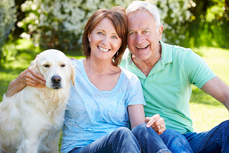 An elderly couple sitting on grass with a golden retriever dog between them, smiling and looking at the camera.