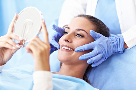 A woman sitting on a dental chair with a blue drape over her shoulders, holding up a magnifying glass over her face, while receiving a dental examination by a dentist wearing gloves.