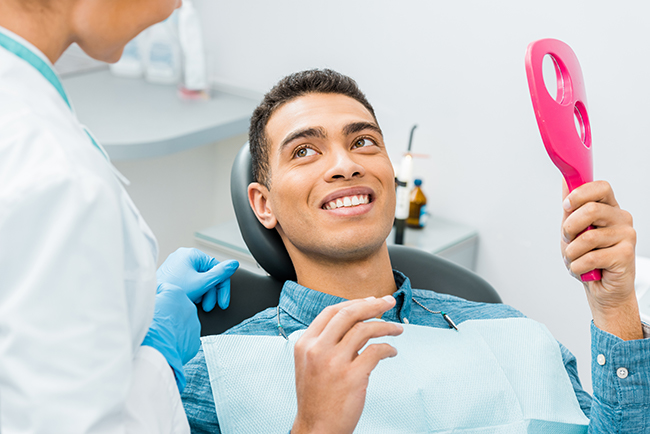 The image shows a person sitting in a dental chair with a smile, holding a pink object that appears to be an oral care device, while being attended by a dental professional who is smiling and wearing a white coat and stethoscope.