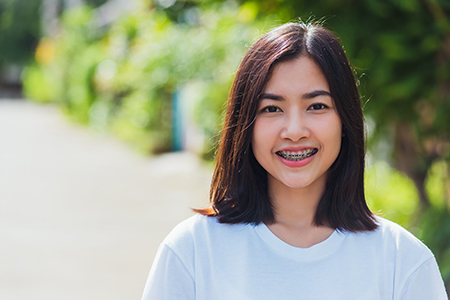The image shows a woman with short hair wearing a white top, smiling at the camera, standing outdoors during daylight.