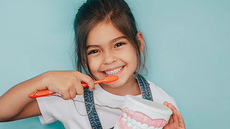 The image features a young girl brushing her teeth with an oversized toothbrush while holding a giant toothpaste tube, with two separate photos side by side showcasing the same activity from different angles.