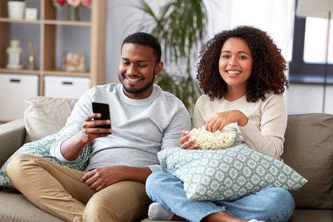 A man and woman sitting on a couch with popcorn, smiling and looking at their phones.