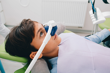 A young person sitting in a dental chair with an instrument over their face, likely for dental examination or treatment.