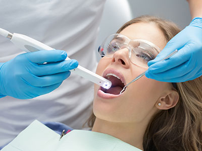 The image shows a woman seated in a dental chair receiving dental care with a device being used on her teeth by a professional wearing protective gloves and a mask, indicating a hygienic and sterile environment.