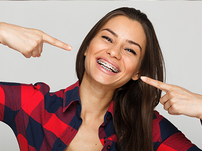 A woman with braces pointing at her teeth with both hands.