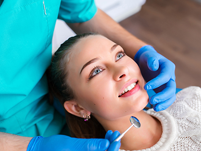The image shows a woman sitting in a dental chair with a dentist performing a procedure on her teeth, wearing blue gloves and using dental instruments.