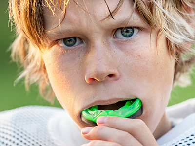 The image shows a young boy with blonde hair holding a green toothbrush in his mouth, appearing in two different states of expression, one neutral and one exaggeratedly surprised, with a football field in the background.