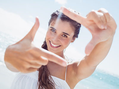 The image shows a smiling woman taking a selfie with her hand pointing towards the camera lens, set against a beach backdrop with clear skies and white clouds.