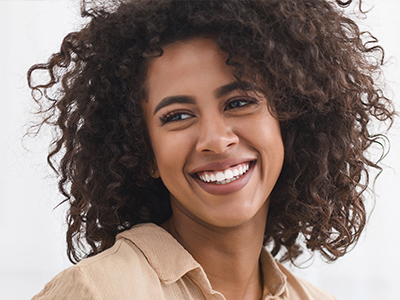 A woman with curly hair smiles brightly at the camera, wearing a beige top and standing against a white background.