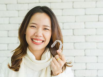 A woman holding a toothbrush smiles at the camera, while another person stands behind her with their mouth closed in a neutral expression.